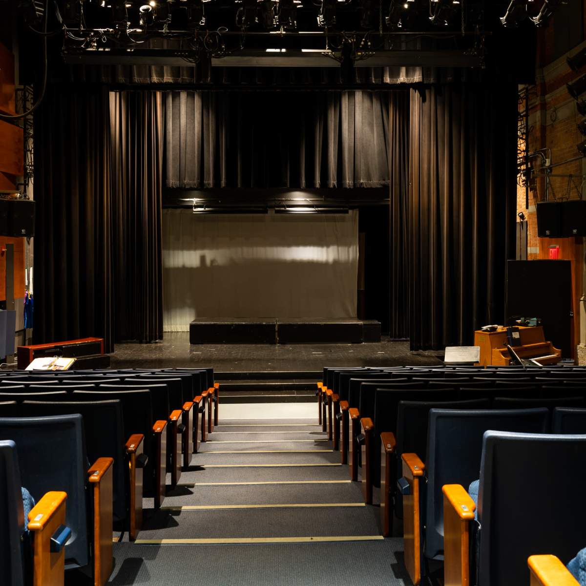 An empty theater with rows of blue seats facing a dark stage draped in black curtains.
