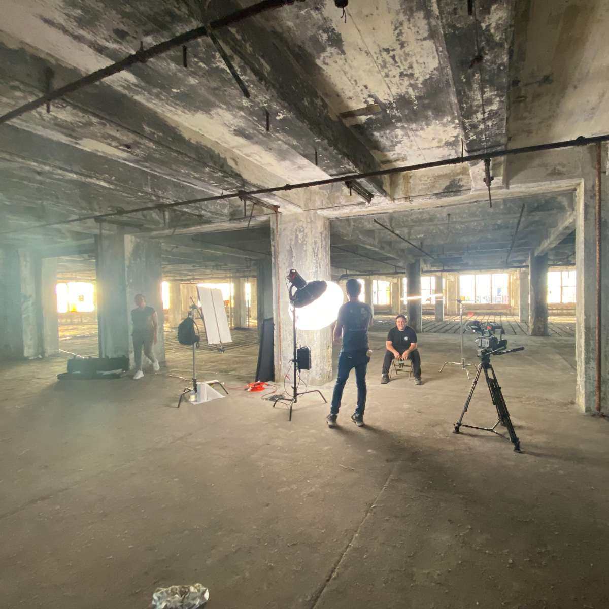 A film crew setting up a shot in a large, industrial, abandoned space with concrete pillars and uneven lighting.