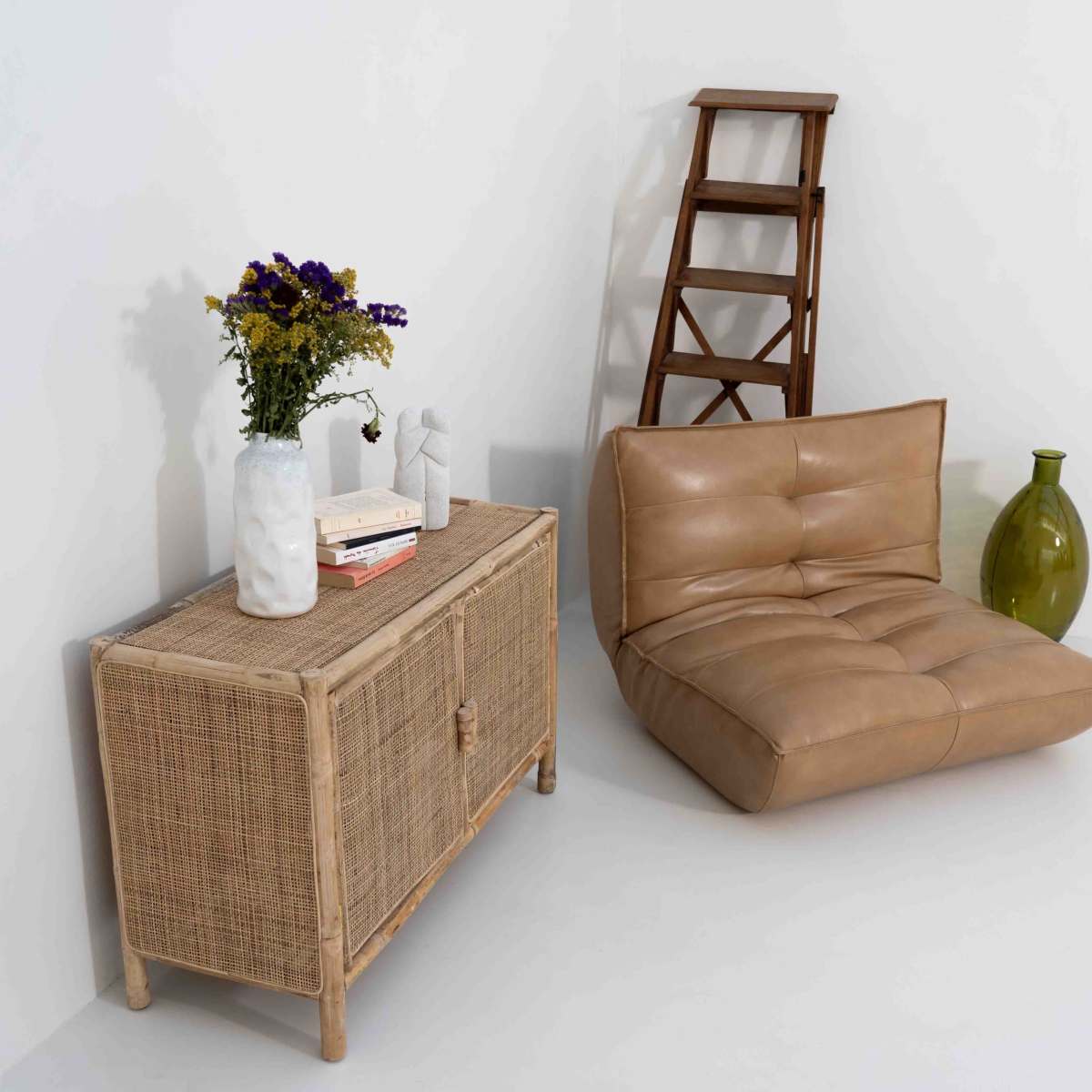 A minimalist interior setting featuring a beige leather chair, a wicker cabinet with a flower vase, and a wooden ladder against a white wall.