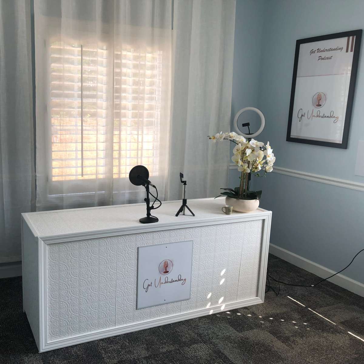 A brightly lit room with a white desk featuring a microphone, a lamp, and an ornamental plant, situated near a window with plantation shutters.