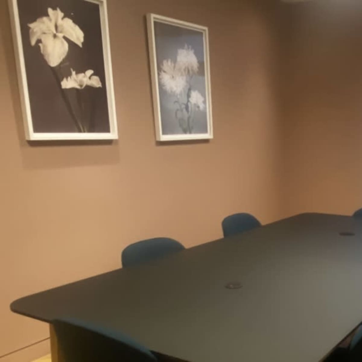 A conference room features a large black table surrounded by four chairs, with two floral artworks on the wall and a window partially covered by a sheer curtain.