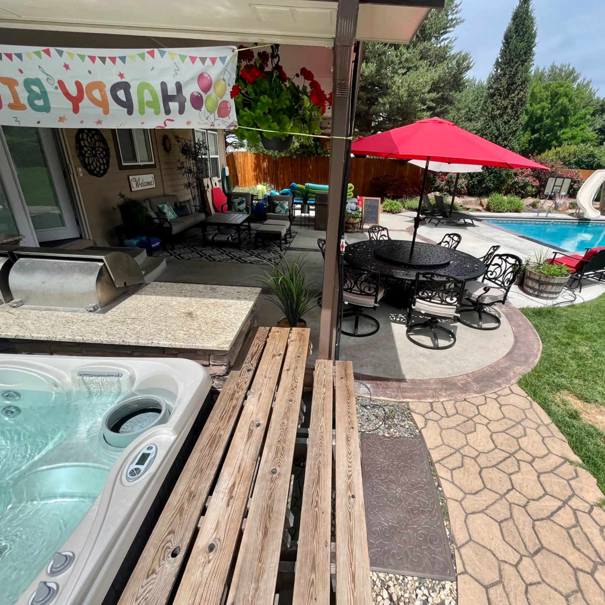 A backyard with a hot tub, a dining area under a canopy, a swimming pool, and a red umbrella beside green grass.
