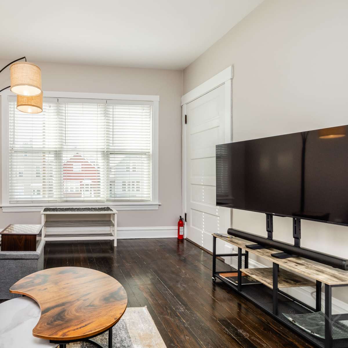 A living room features a gray sofa, a floor lamp with two shades, a coffee table, a television stand, and a window with white blinds.