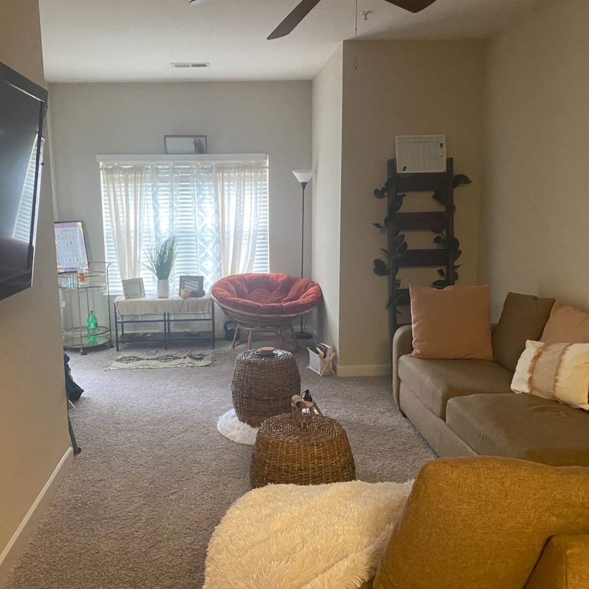 A living room with a large television, a sectional sofa, a round chair, and two woven ottomans.