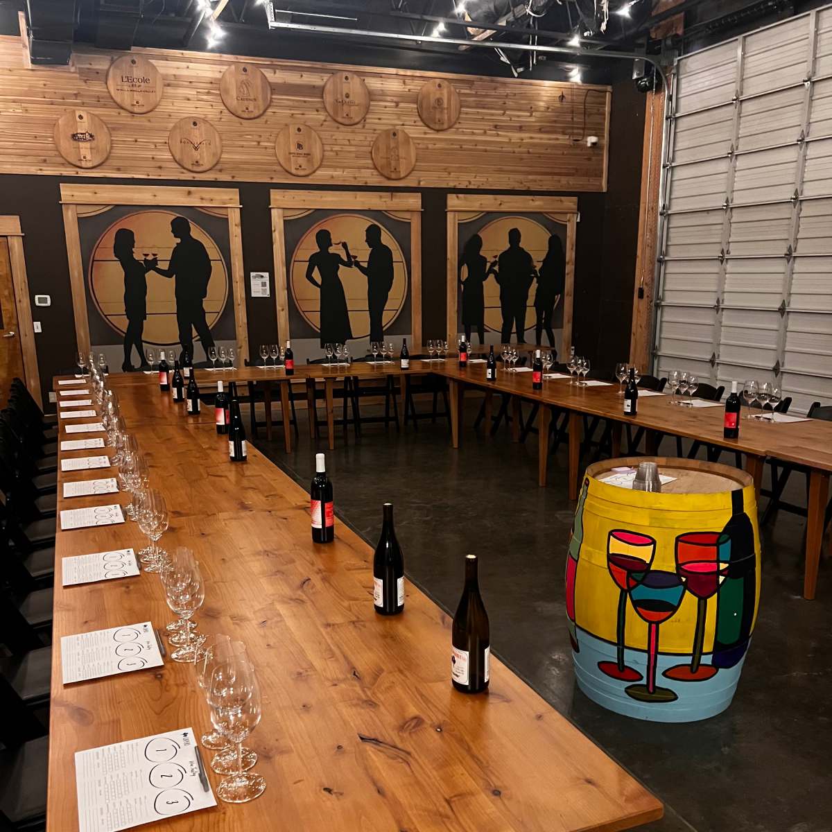 A large wooden dinner table is arranged in a U-shape with wine glasses and bottles, surrounded by silhouetted figures on the walls and a colorful painted barrel at one end.