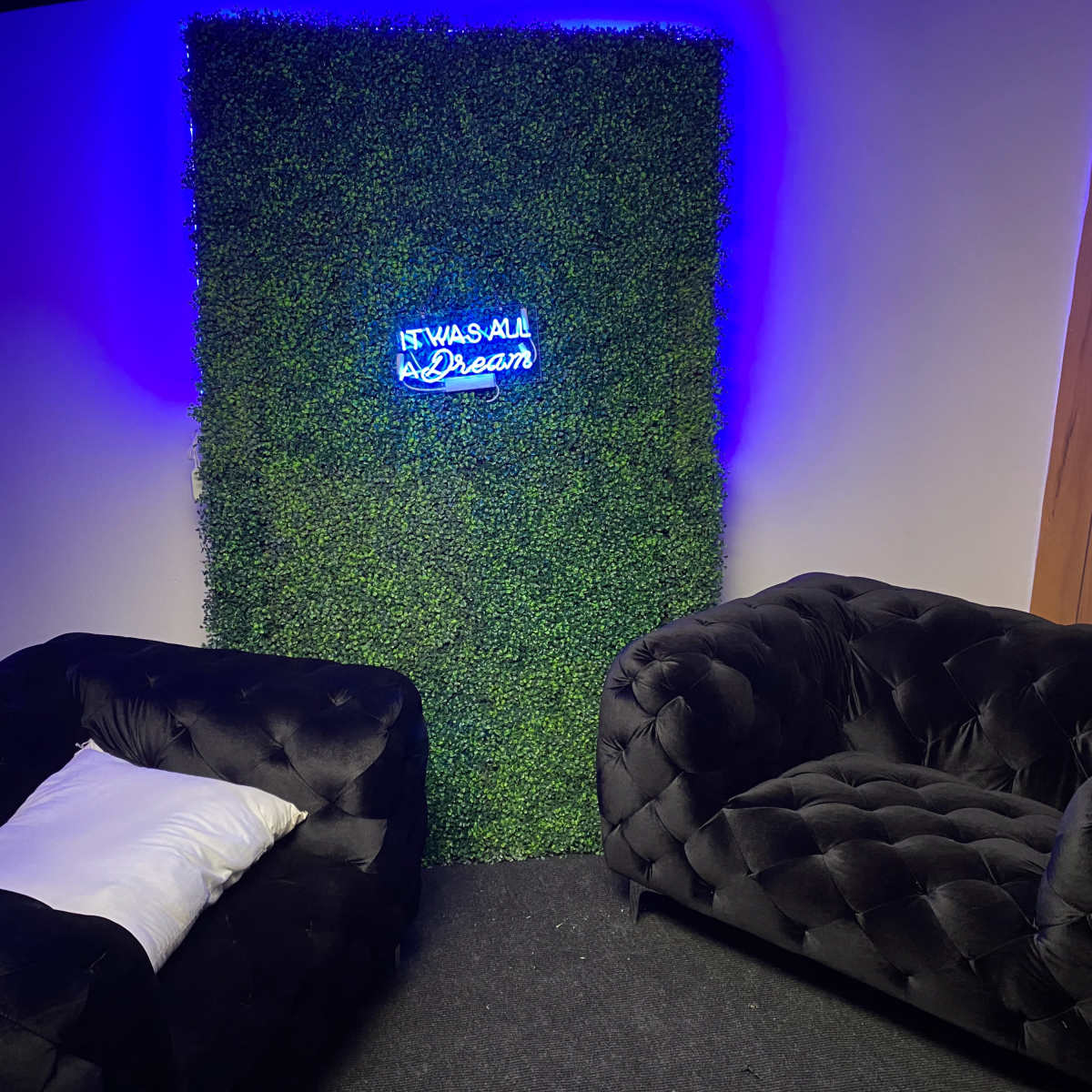 A small room features two black tufted sofas facing a green wall adorned with a neon sign that reads, "It Was All a Dream."