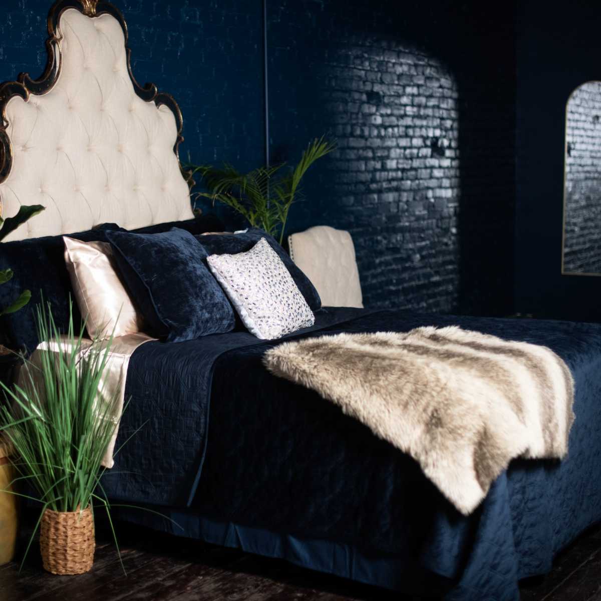 A elegantly styled bedroom featuring a tufted headboard, dark blue walls, and a bed adorned with navy bedding and decorative pillows, alongside potted plants and a fur throw.