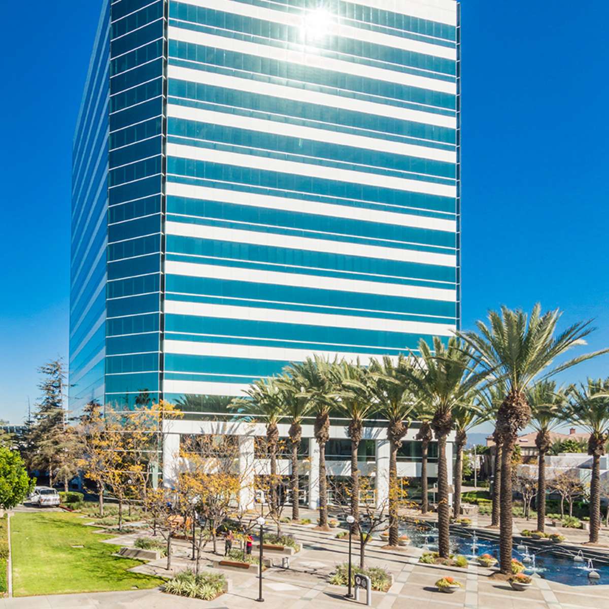 A modern glass office building stands tall next to a row of palm trees and landscaped walkways under a clear blue sky.