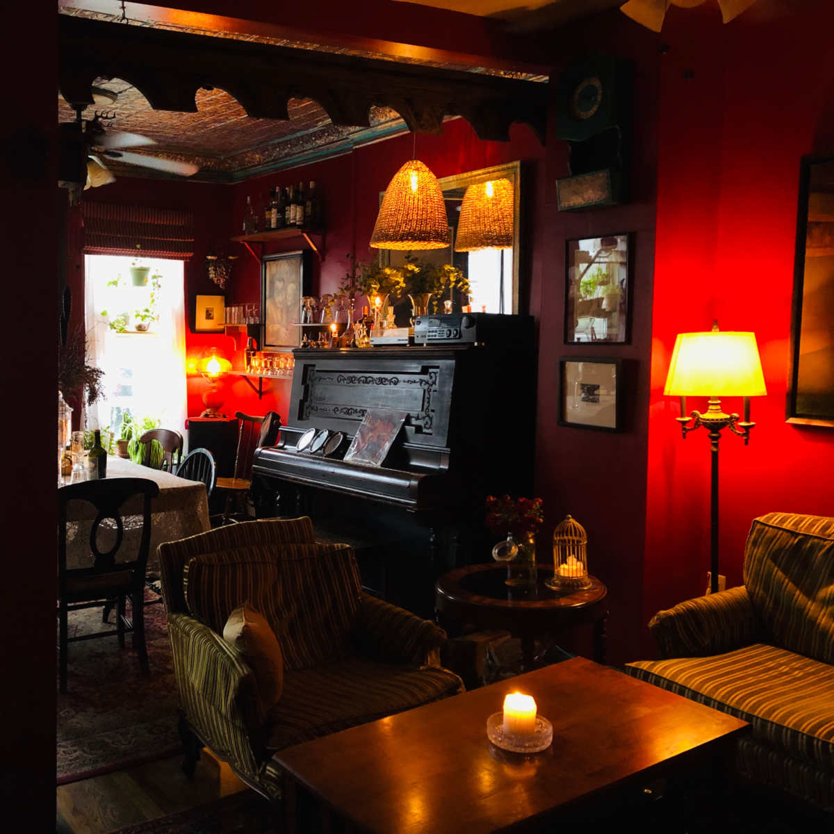 A cozy lounge area features a piano, vintage furniture, and warm lighting, with plants visible through large windows.