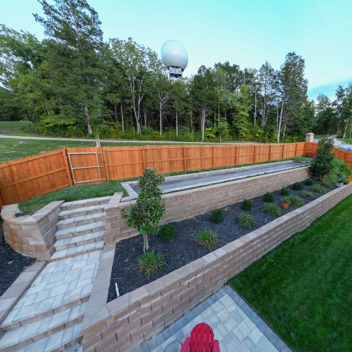 A landscaped backyard with a stone staircase leading to a raised garden bed bordered by wooden fencing and trees in the background.