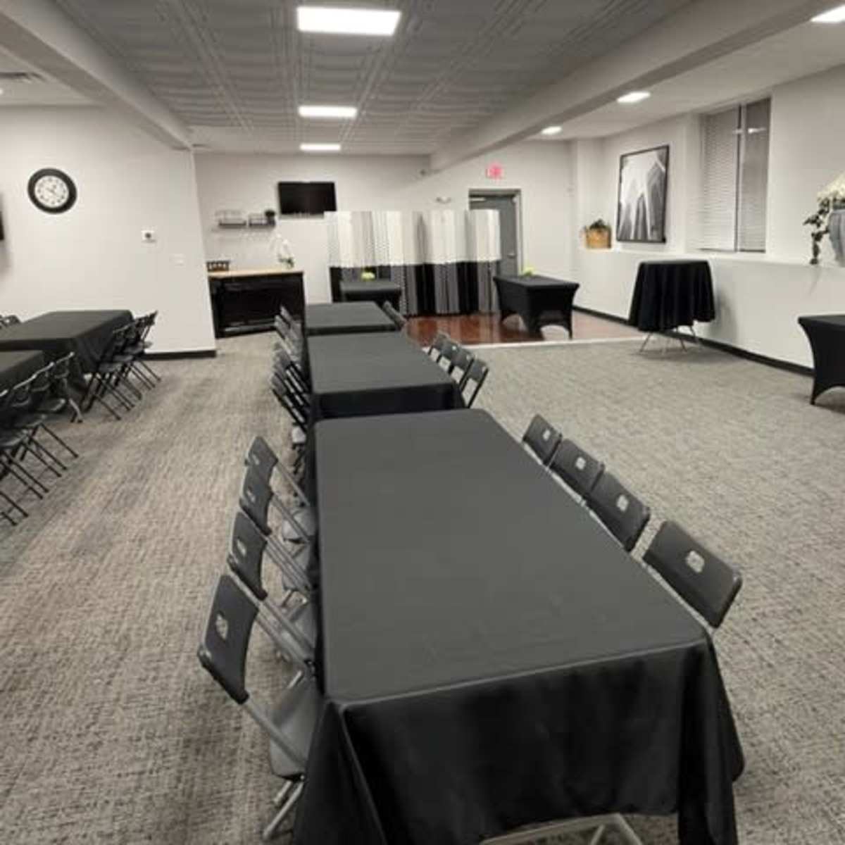 A spacious meeting room with multiple long tables covered in black cloth, surrounded by folding chairs, and equipped with a kitchenette area in the background.