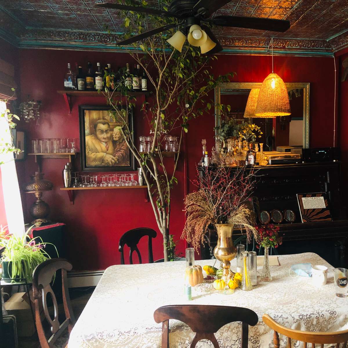 A cozy dining area with a round table set with a lace tablecloth, surrounded by wooden chairs, and featuring a piano in the background along with various plants and vintage decor.