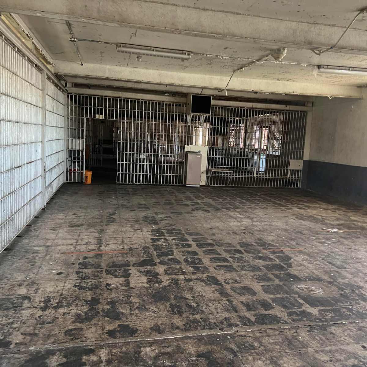 A large, empty room with concrete flooring and barred windows, featuring visible signs of wear on the walls and floor.