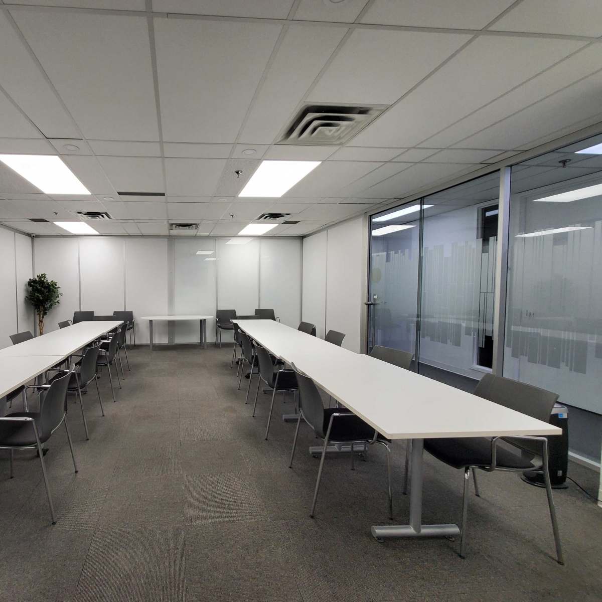 A spacious conference room features two long tables arranged in a U-shape, surrounded by chairs and fitted with glass walls and a projector screen at one end.