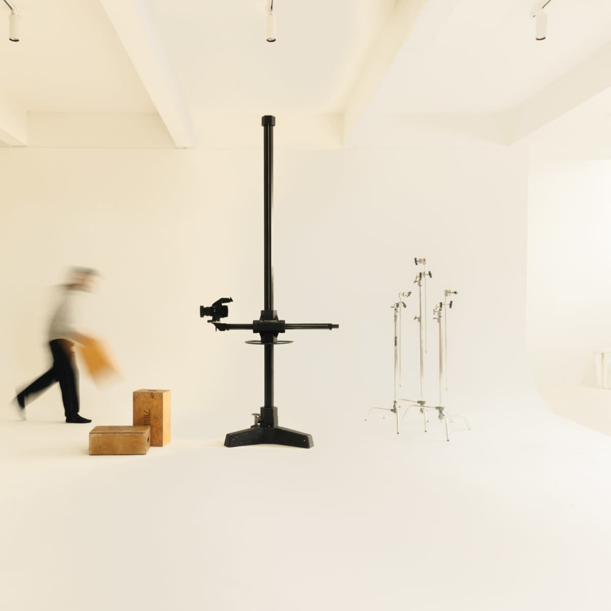 A person carrying an item walks past a large camera stand in a minimalist studio space.