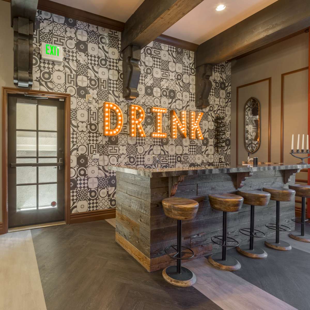A stylish bar area with a wooden counter and stools, illuminated with the word "DRINK" made of bright lights on a patterned wall.