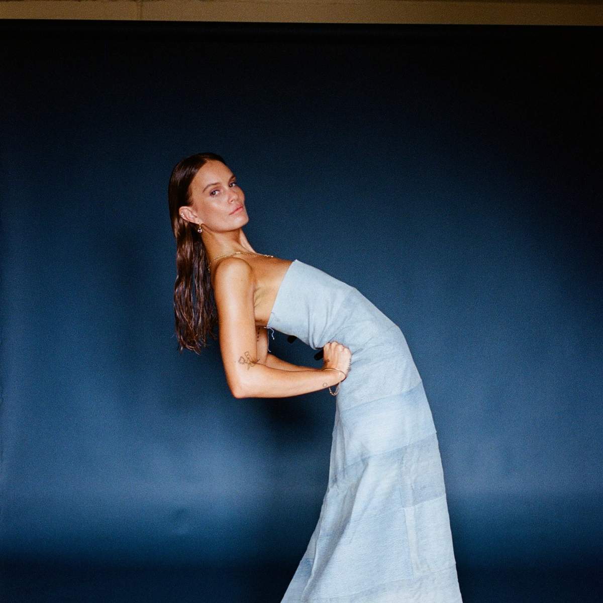 A model in a fitted, floor-length dress poses against a dark blue backdrop.