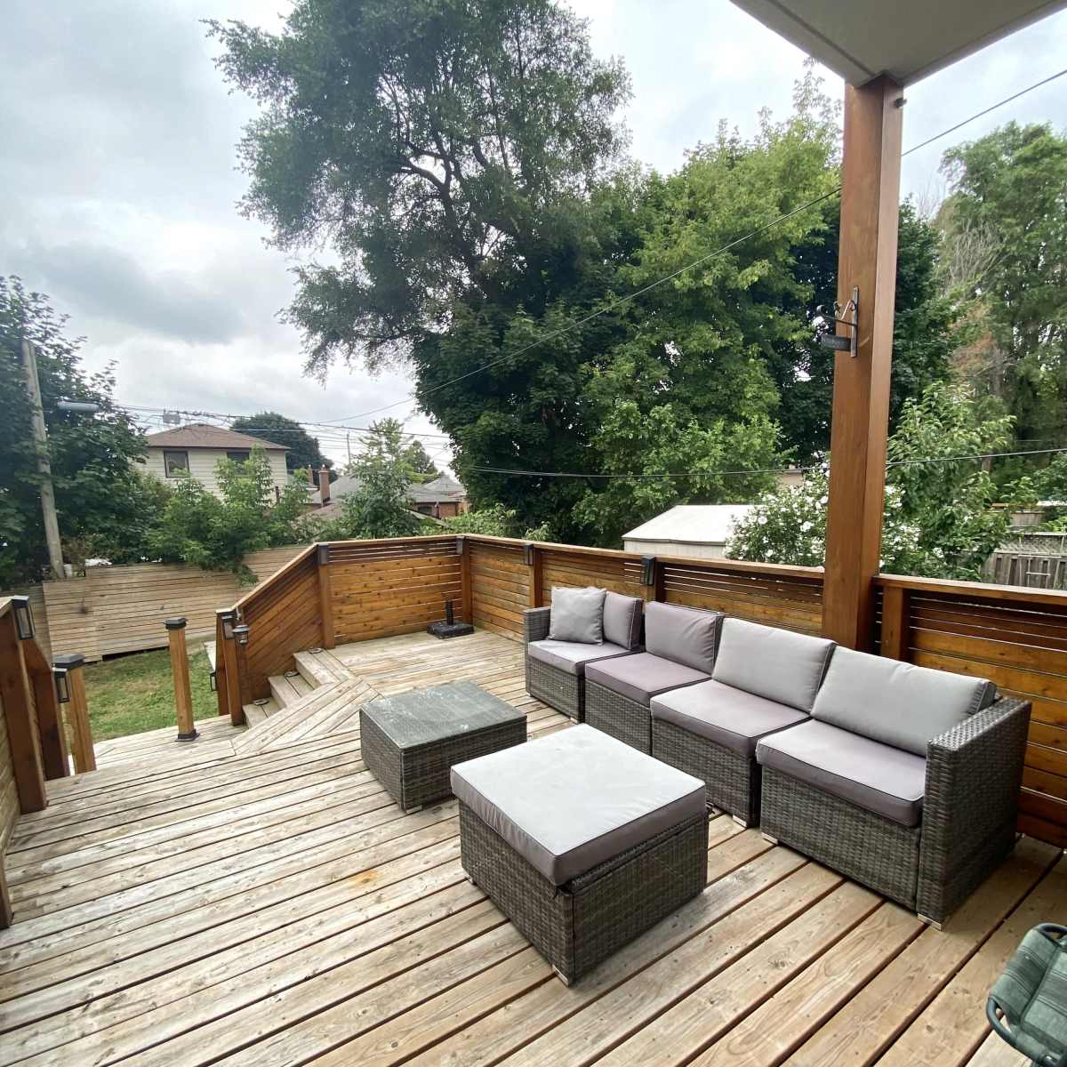 A wooden deck features a seating area with gray wicker furniture and a glass table, surrounded by trees and a grassy yard.
