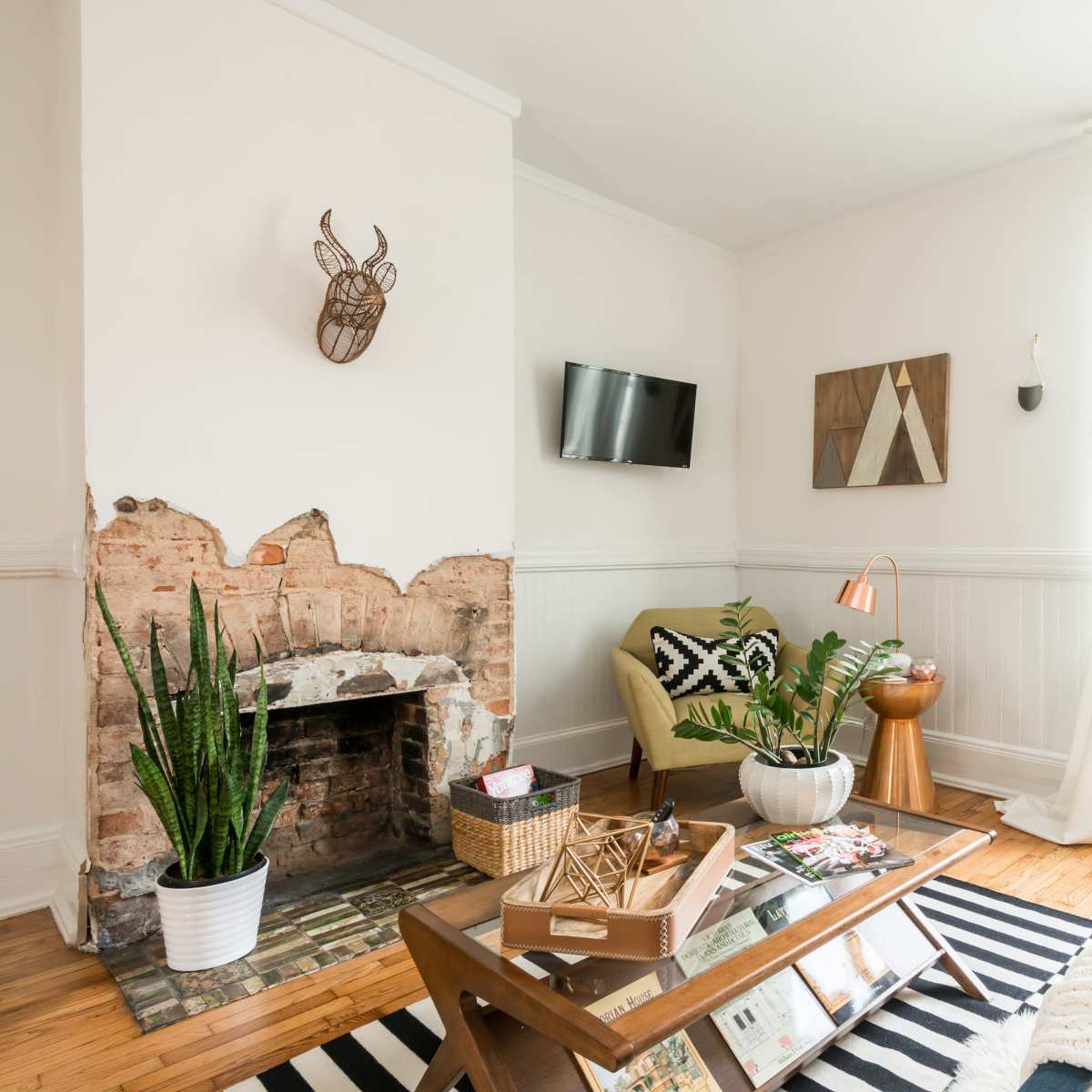 A bright living room featuring a mounted decorative mask, a small flat-screen TV, a fireplace with exposed brick, and various furniture pieces, including a green armchair and a coffee table with books and magazines.