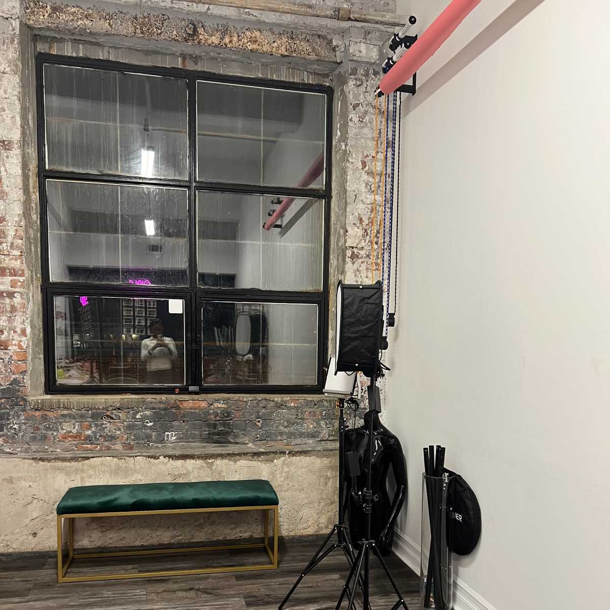 A corner of a room featuring a large window with a black frame, a green bench, and a light stand beside various black equipment.