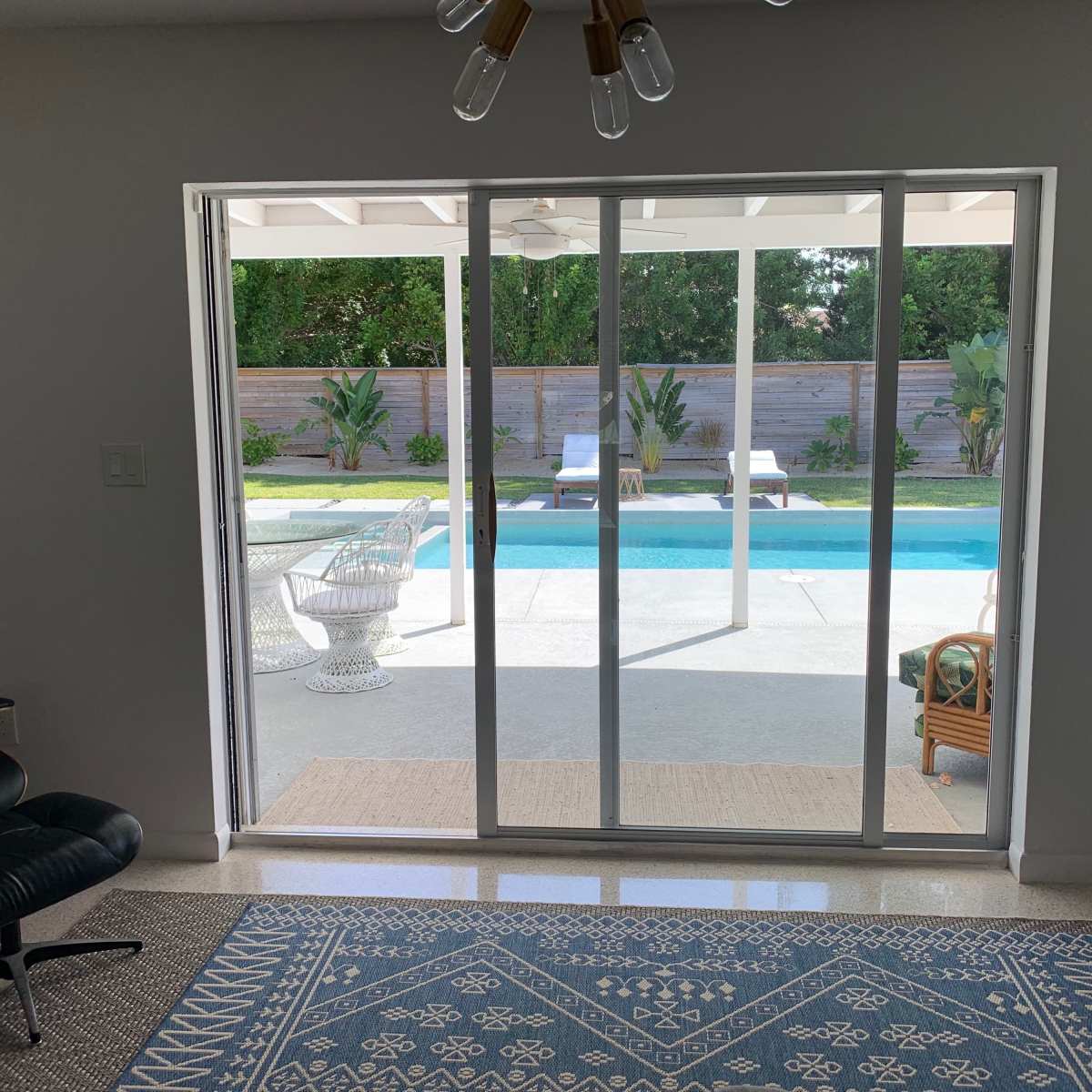A living room with a sliding glass door that opens to a backyard featuring a pool, lounge chairs, and landscaped greenery.