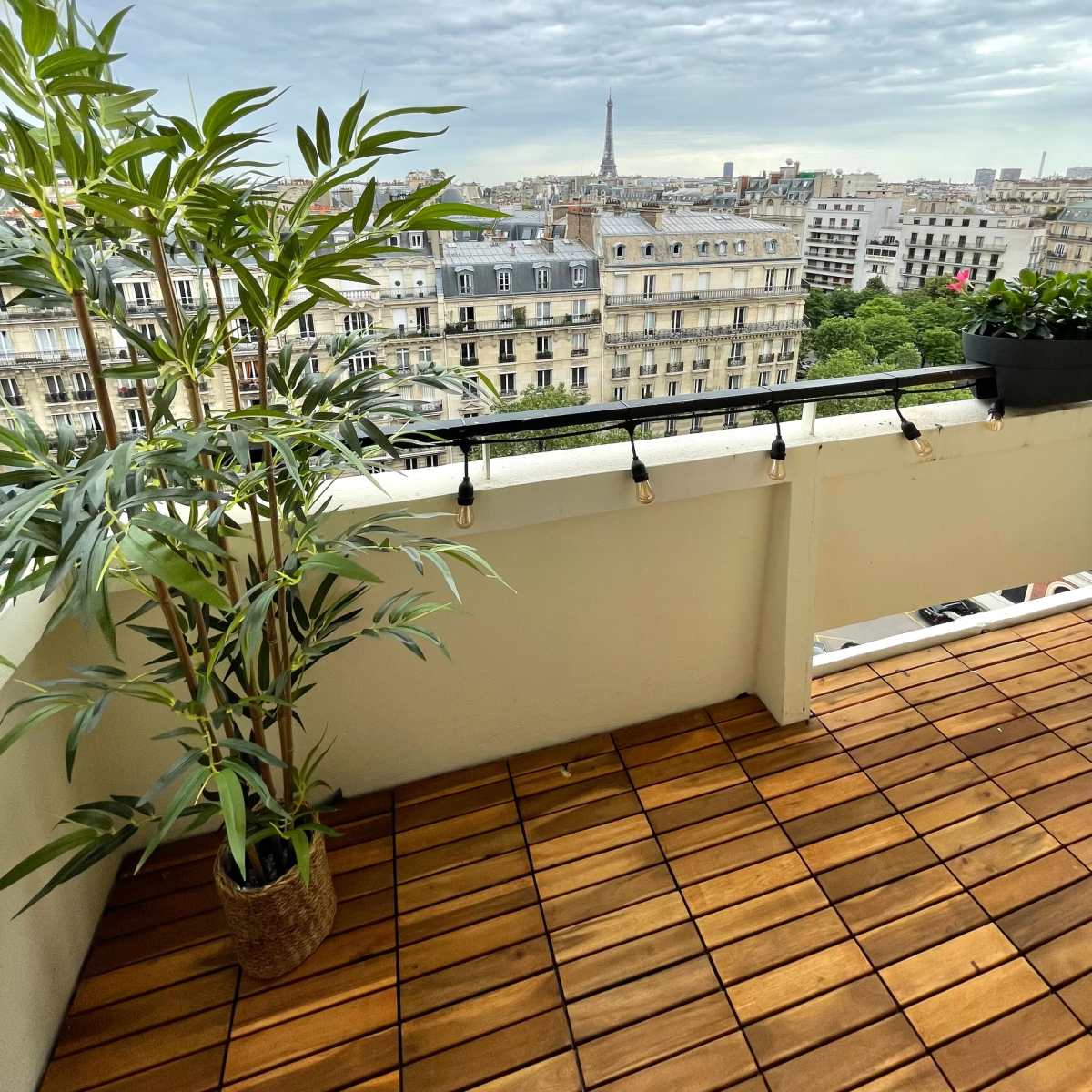 A balcony with wooden flooring features a potted plant and offers a view of Paris, including the Eiffel Tower in the distance under a cloudy sky.