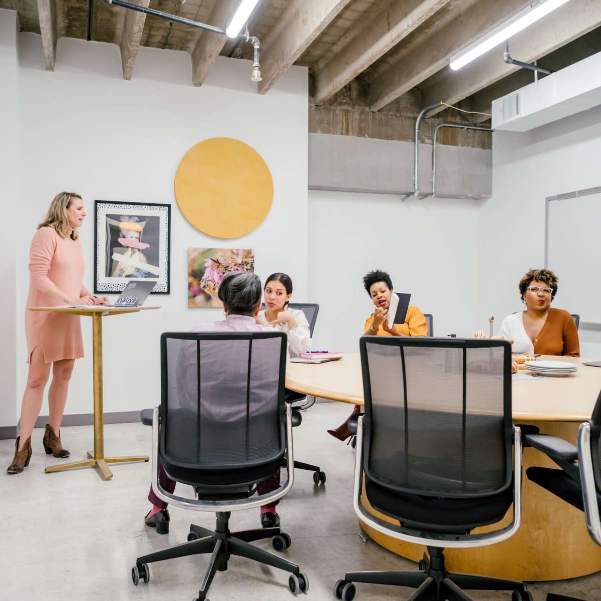 A group of people engage in a meeting around a large table in a modern conference room, while a woman stands at a nearby podium presenting.