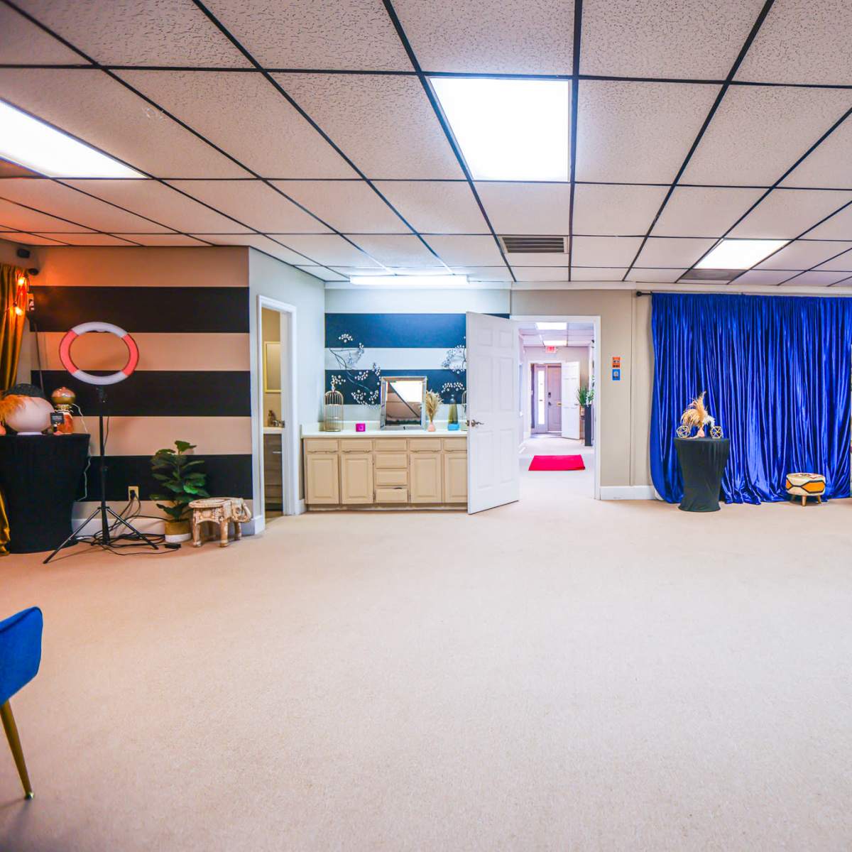 A spacious room with beige carpet, a visible entrance leading to a red carpet hallway, and blue curtains on one side, along with various furniture and decorations.
