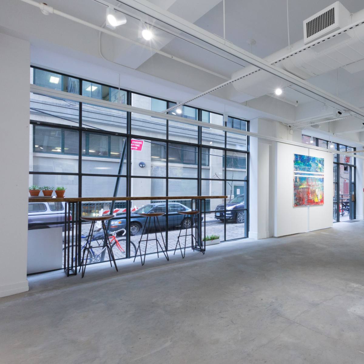 A spacious art gallery with large windows, concrete flooring, and various artworks displayed on the walls.