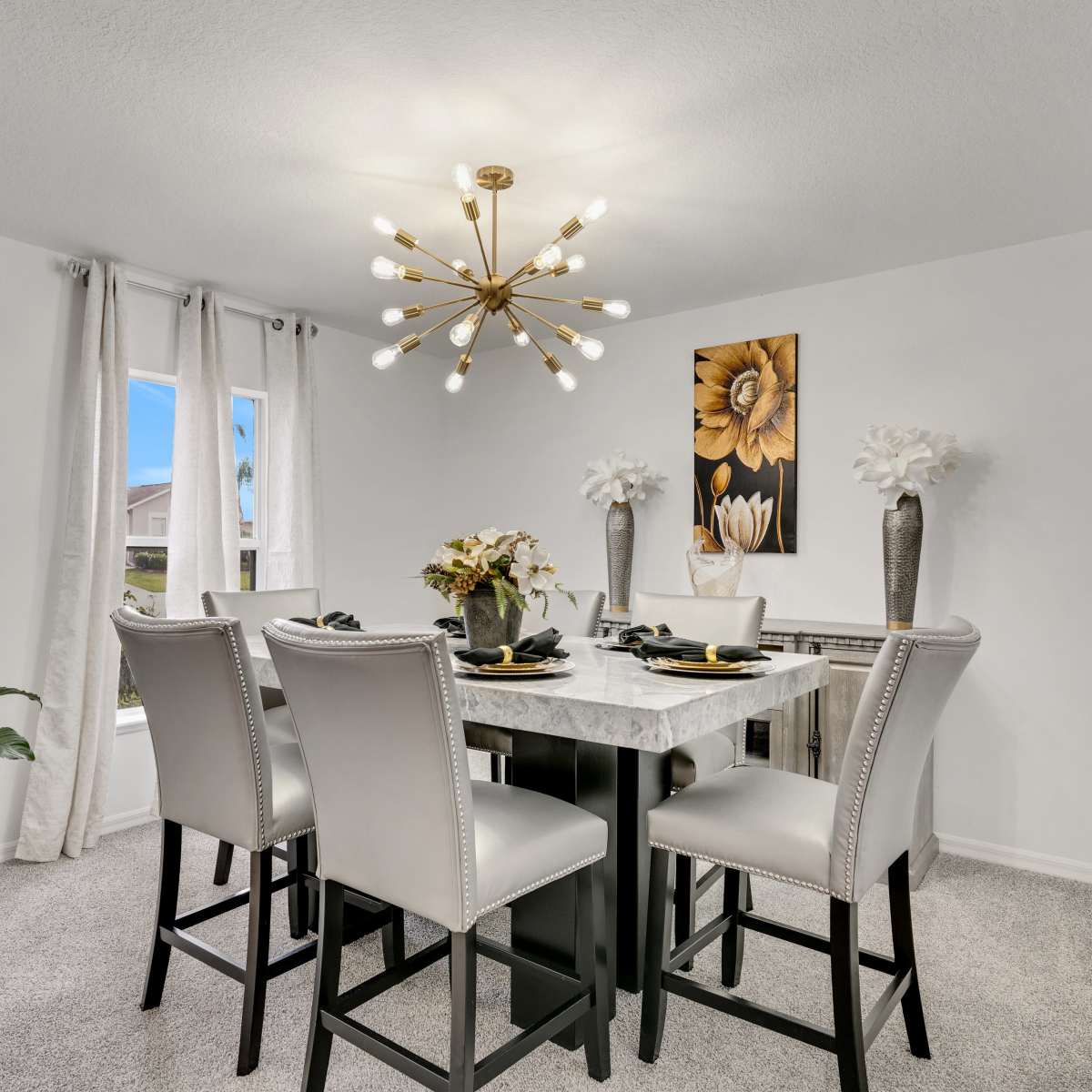 A modern dining area features a marble-top table surrounded by four upholstered chairs, illuminated by a chandelier, with decorative plants and artwork on the walls.