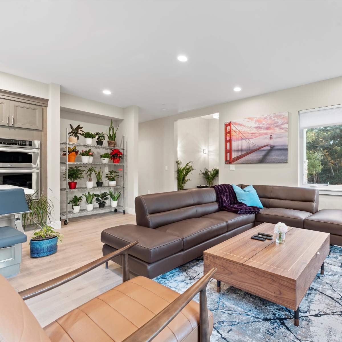 A modern living room with a sectional couch, a wooden coffee table, a large window featuring greenery outside, and decorative plants on shelves.