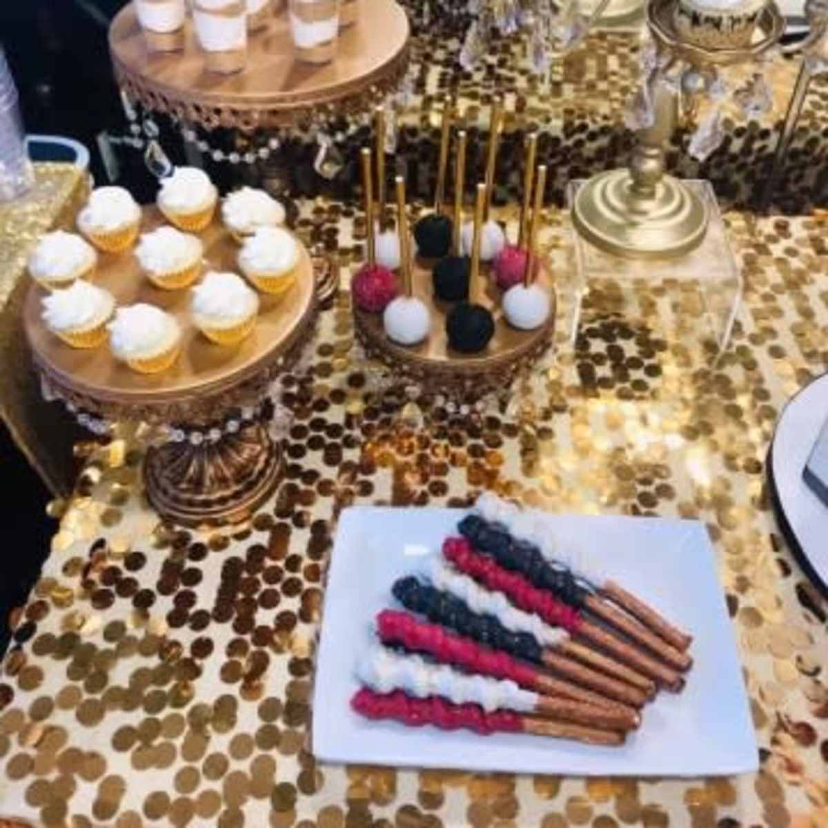 A dessert table featuring various styled cupcakes, mini desserts in cups, and decorated chocolate-dipped pretzel sticks arranged on golden and silver platters.