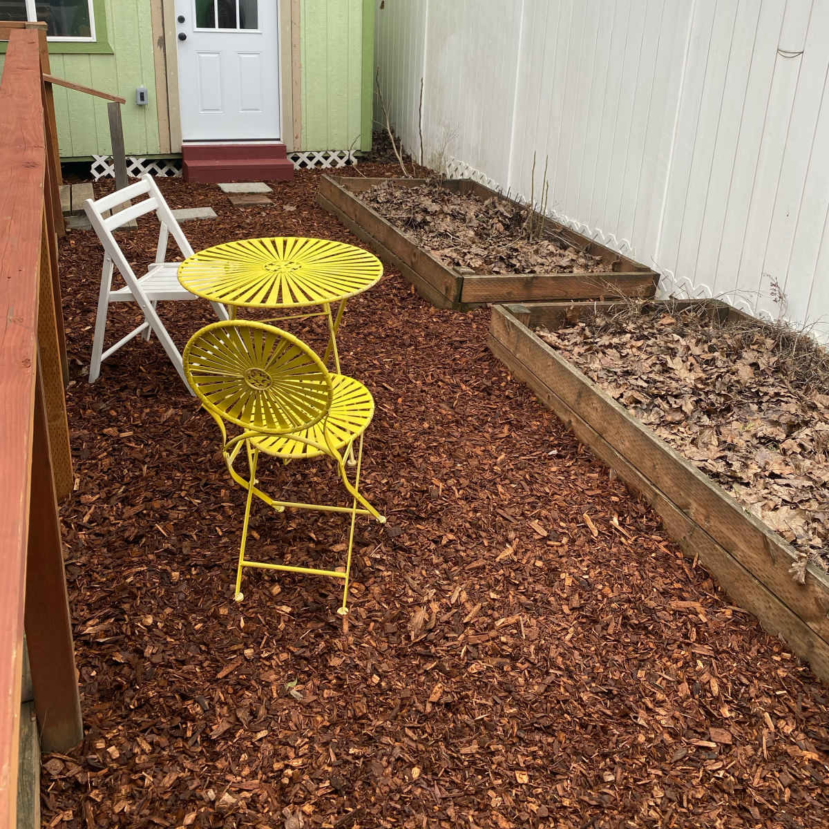 A small patio area features a yellow bistro table and chairs surrounded by wooden garden beds and a white fence.