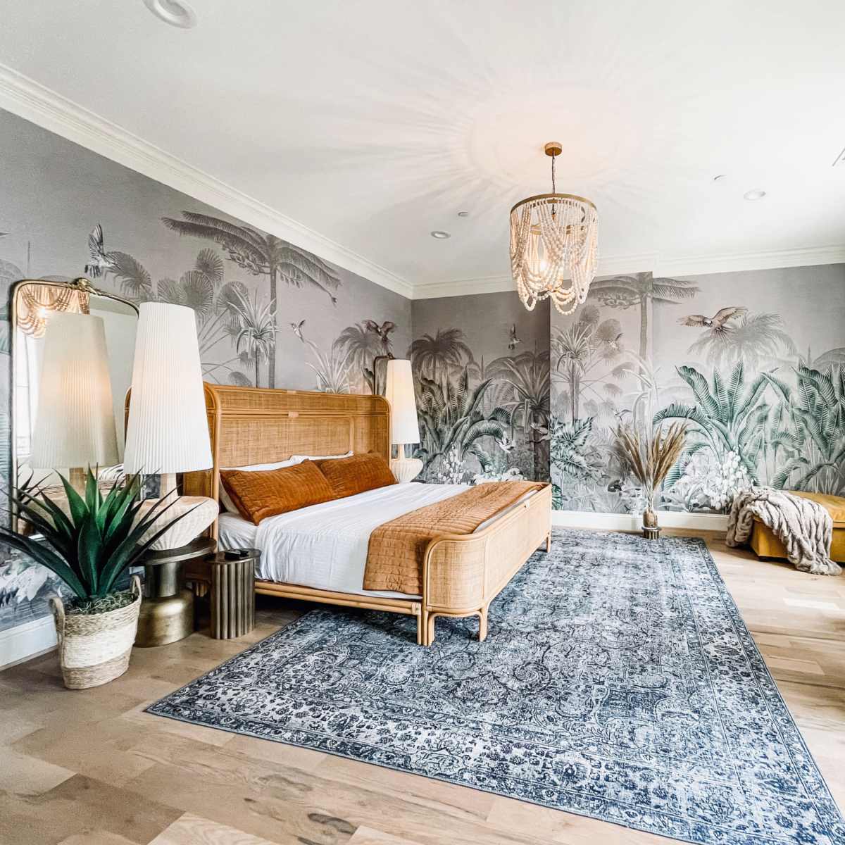 A spacious bedroom features a rattan bed, tropical mural walls, decorative plants, and a woven area rug.