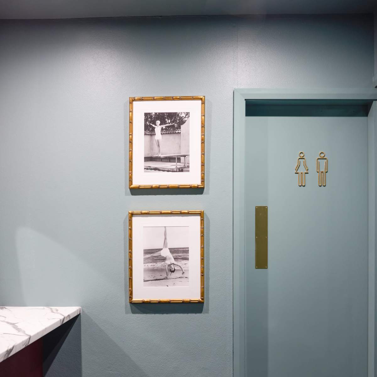 A hallway features a teal wall with two framed black-and-white photographs above a round blue ottoman and a nearby restroom sign next to an elevator door.