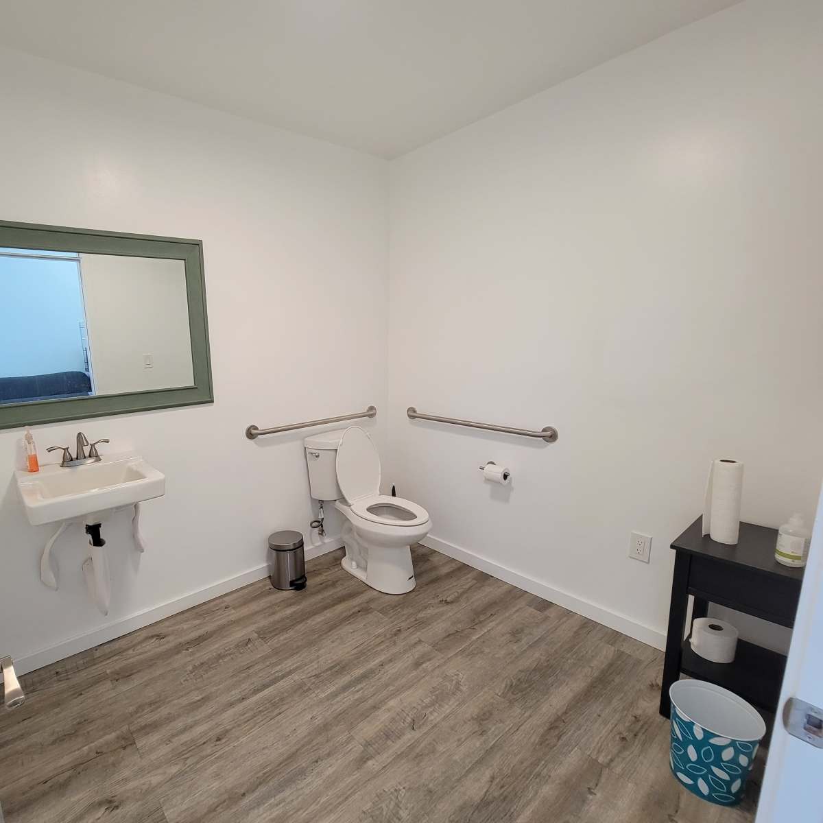 A clean, modern bathroom with a sink, toilet, and a large wall mirror, featuring grab bars and a simple storage table.
