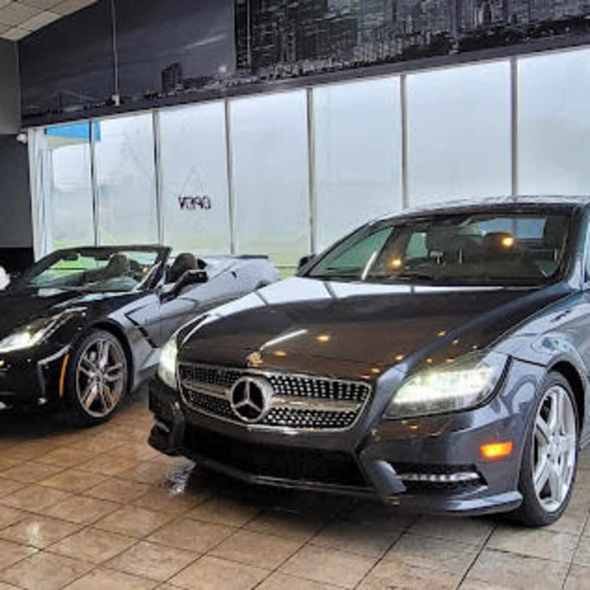 Three luxury cars, including a white Mercedes, a black convertible, and a black Mercedes, are parked inside a storefront with large windows.