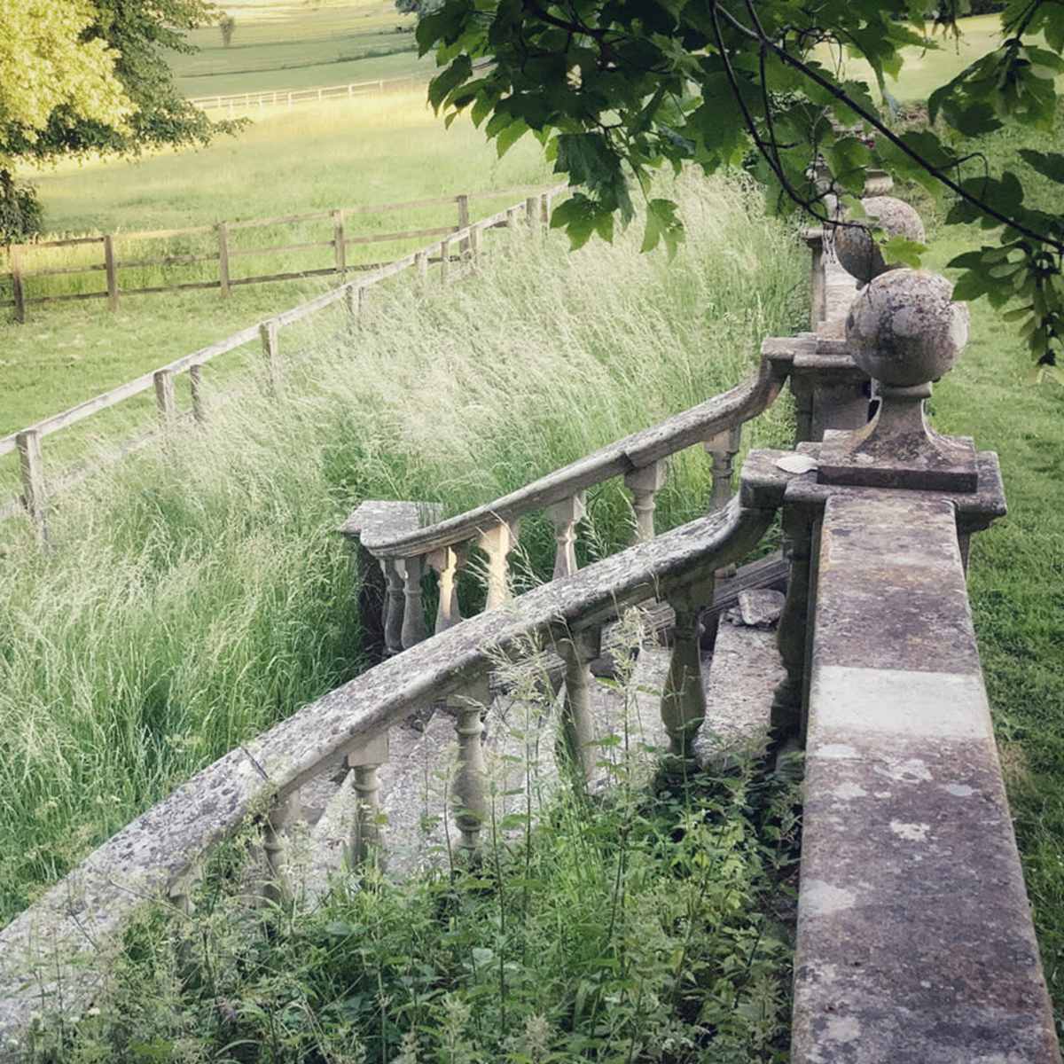A stone balustrade curves alongside a field overgrown with tall grass and framed by trees.