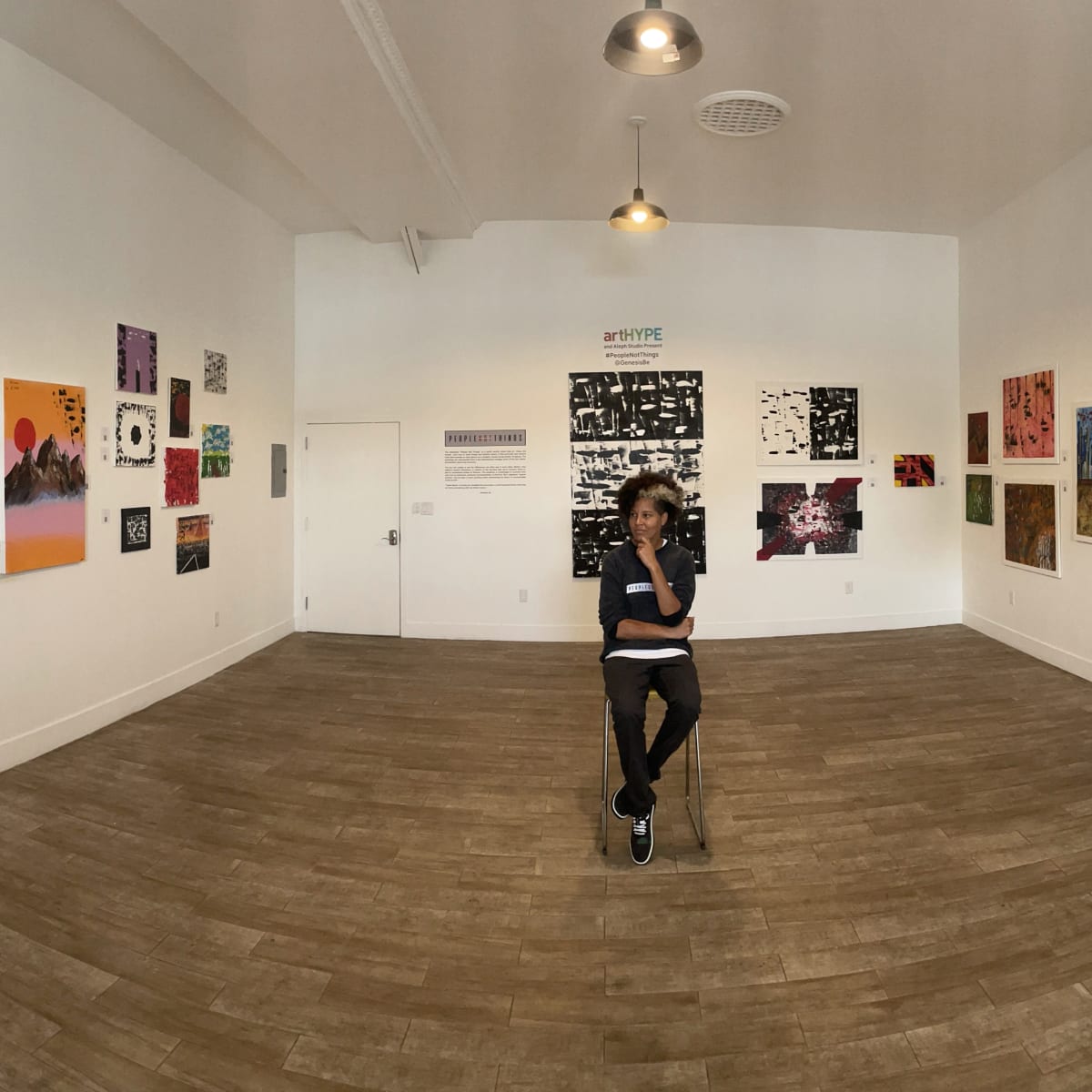 A person sits on a chair in the center of a gallery filled with various artworks on the walls.