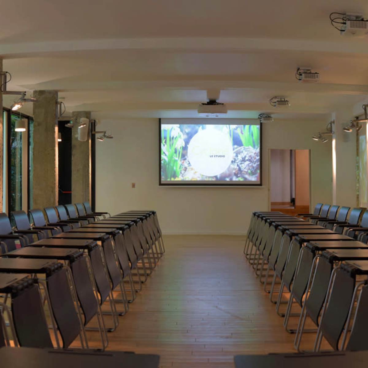 A conference room with rows of black chairs and small tables facing two projection screens, surrounded by large windows that let in natural light.