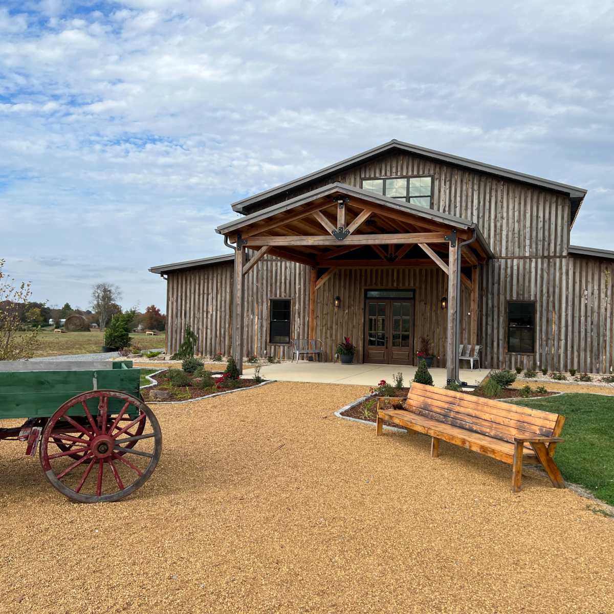 A wooden barn-style building with a covered porch is flanked by a wagon and a wooden bench on a gravel pathway.