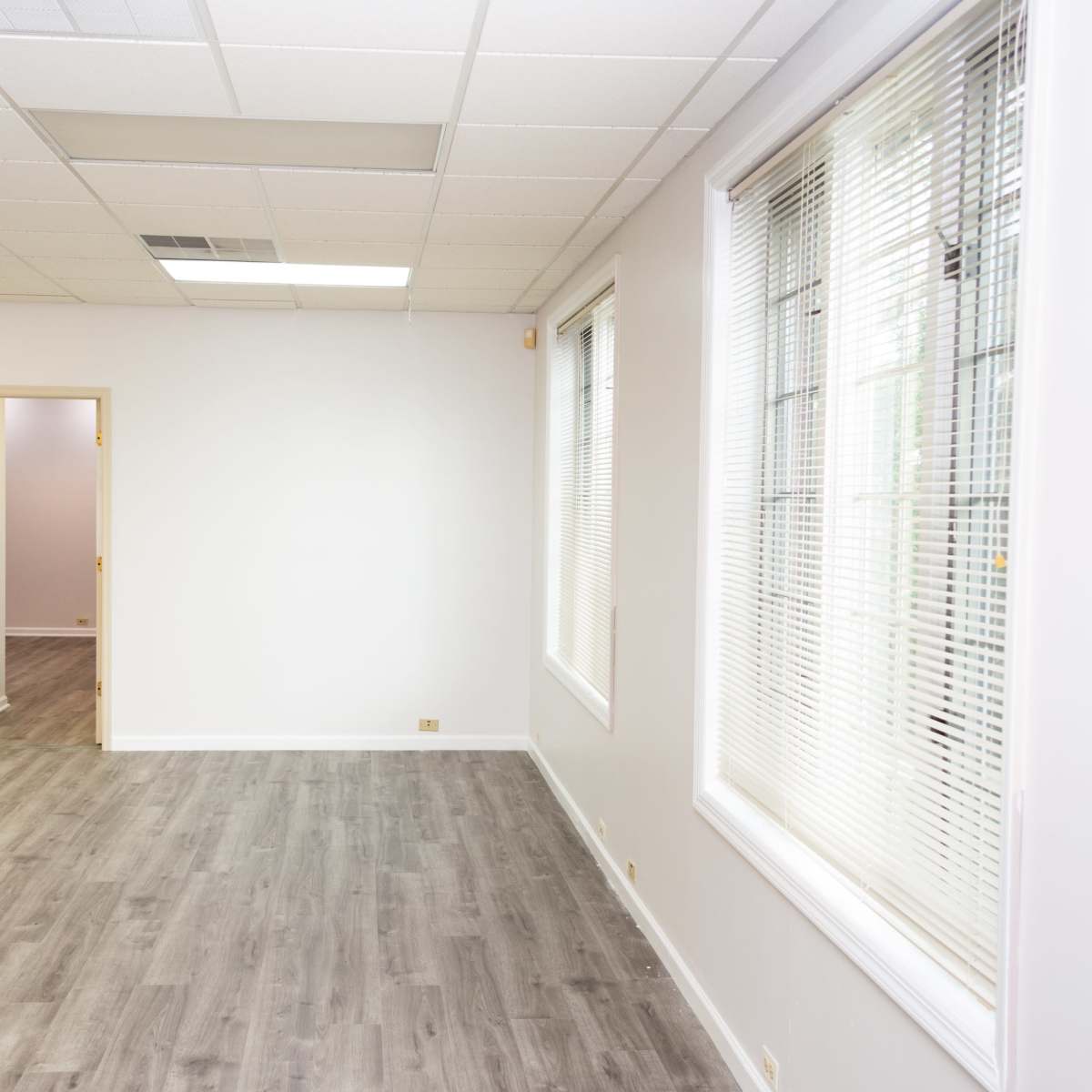 A spacious, unfurnished room with gray flooring, large windows, and a doorway leading to another room.