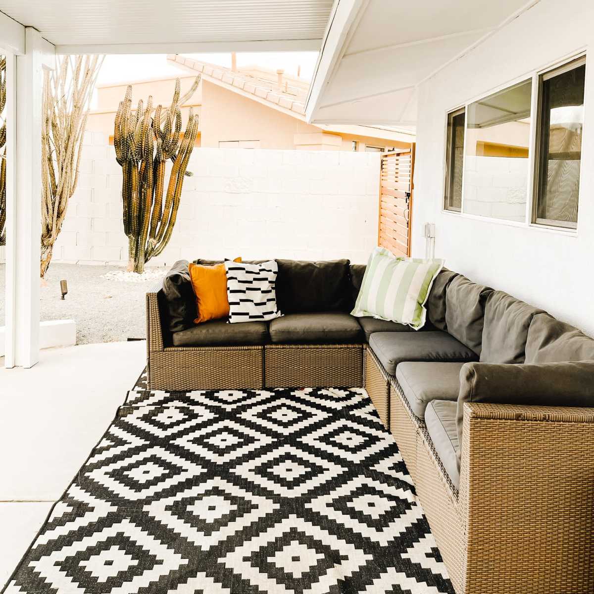 The scene features a patio area with a sectional sofa, a black and white patterned rug, and a backdrop of a low wall and tall cacti.