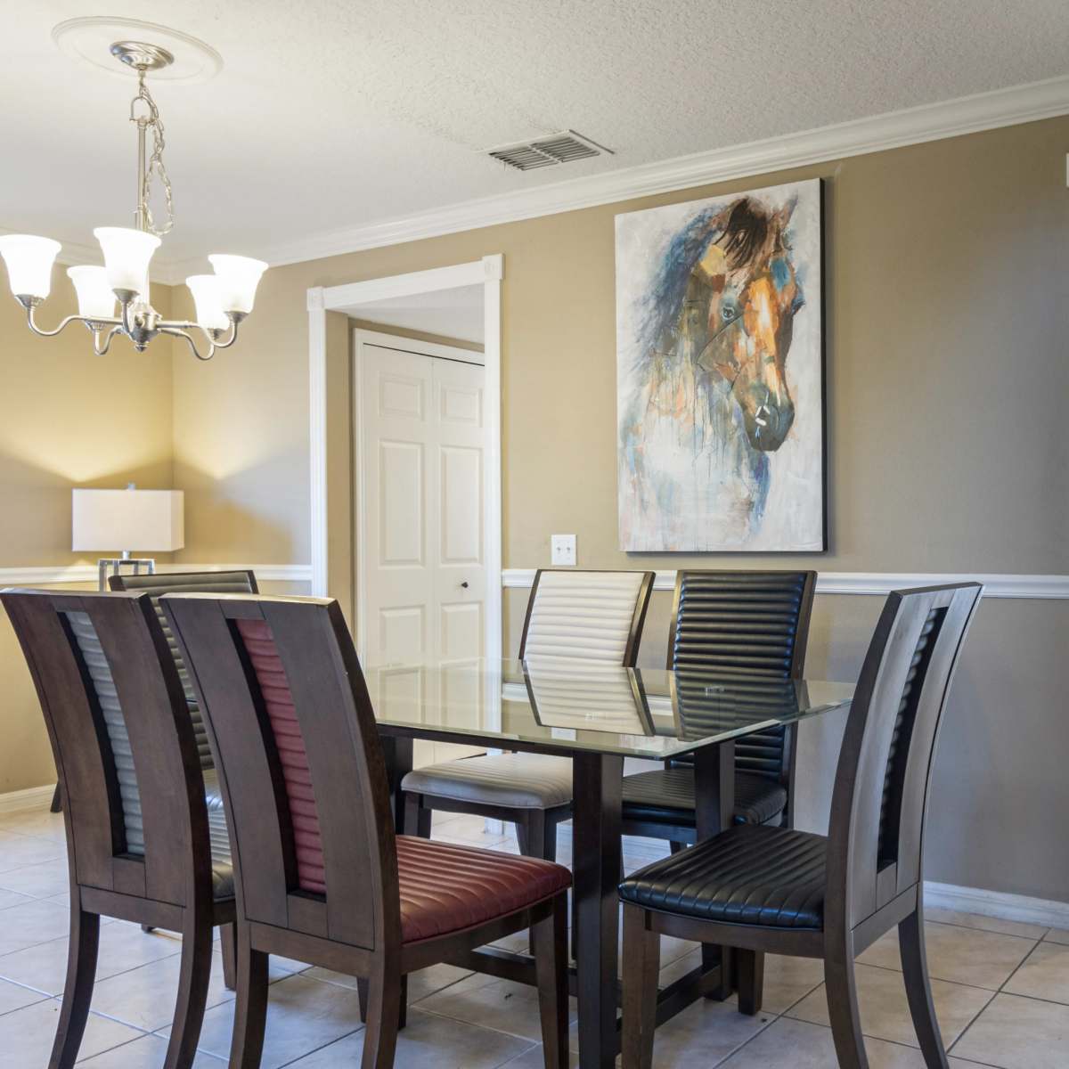 A glass dining table surrounded by wooden chairs is positioned in a well-lit room featuring a painting on the wall and a doorway leading to another space.