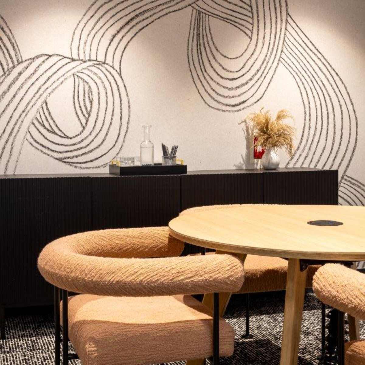 A modern interior featuring a round wooden table surrounded by upholstered chairs, with a decorative wall displaying abstract line art and a sideboard with decorative items.