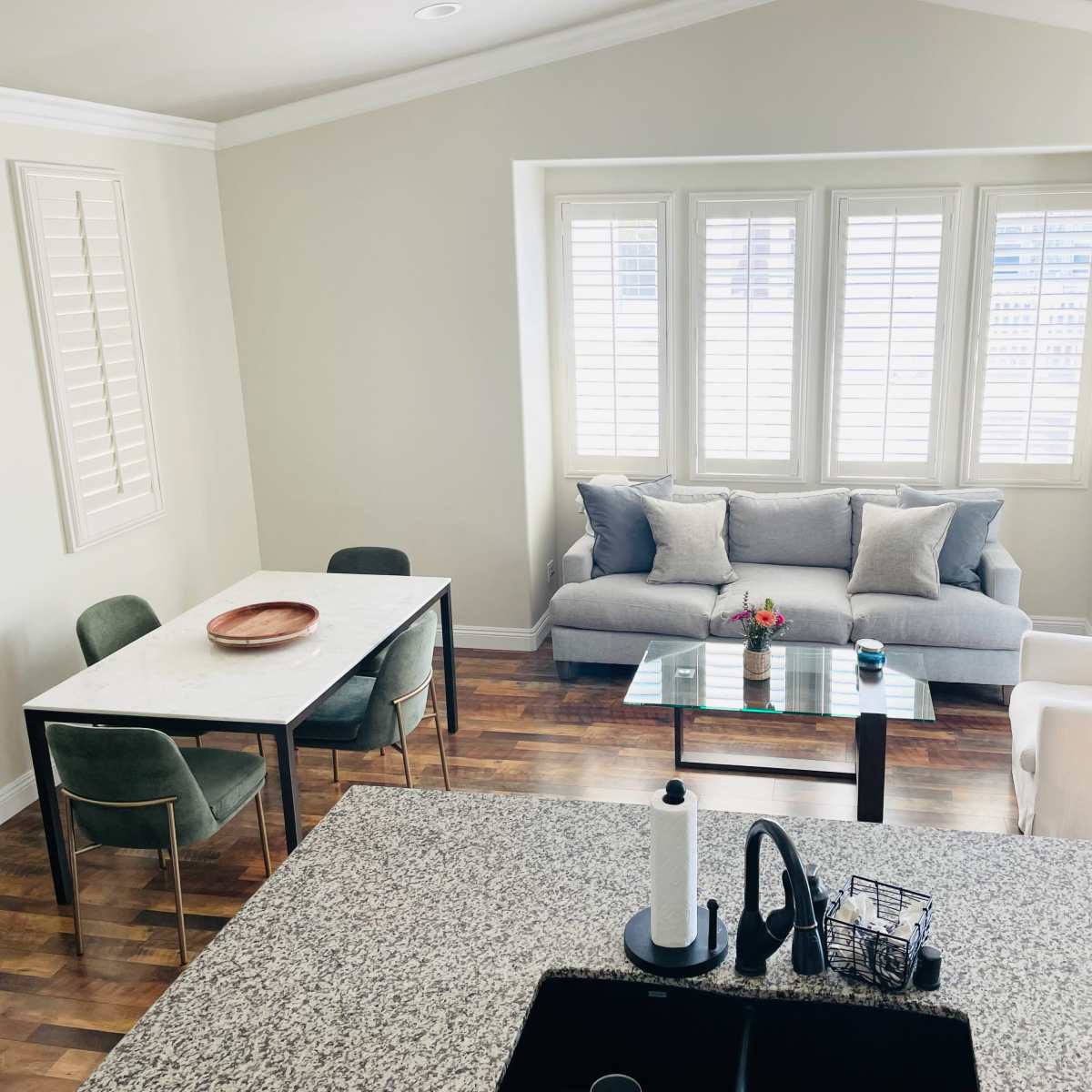 A modern living space features a dining table with chairs, a sofa, and large windows with shutters, all set against a hardwood floor.