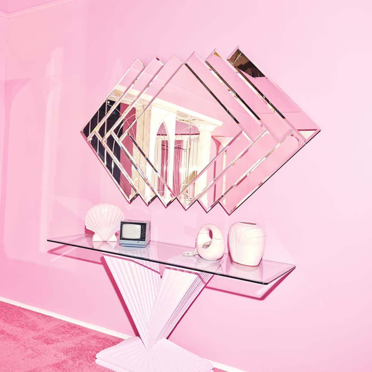 A modern white console table with a geometric design is positioned against a pink wall, featuring a diamond-shaped mirror arrangement above it and various decorative items on the table.