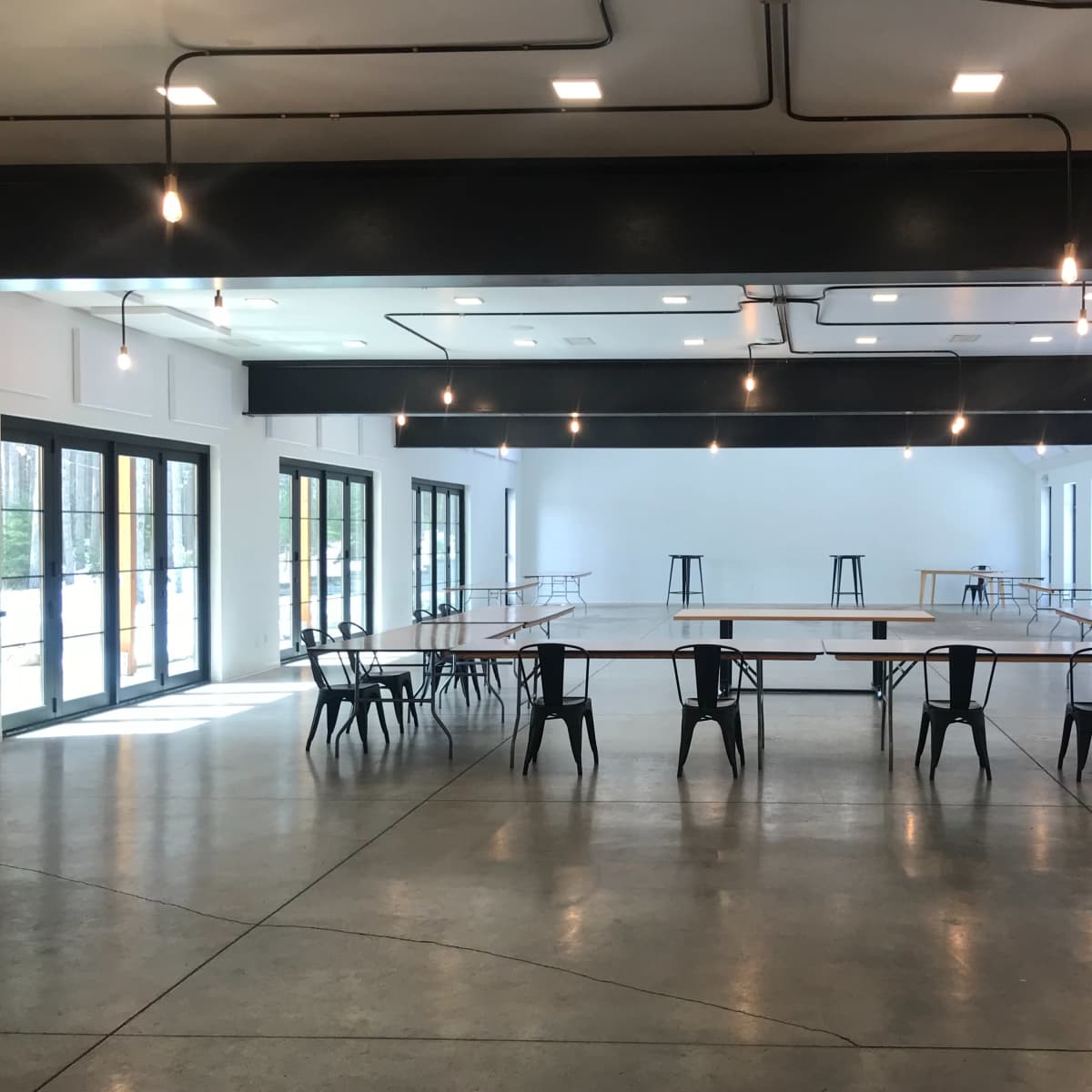 An empty, modern room with several tables arranged in a grid, surrounded by black chairs, and well-lit by hanging lights.