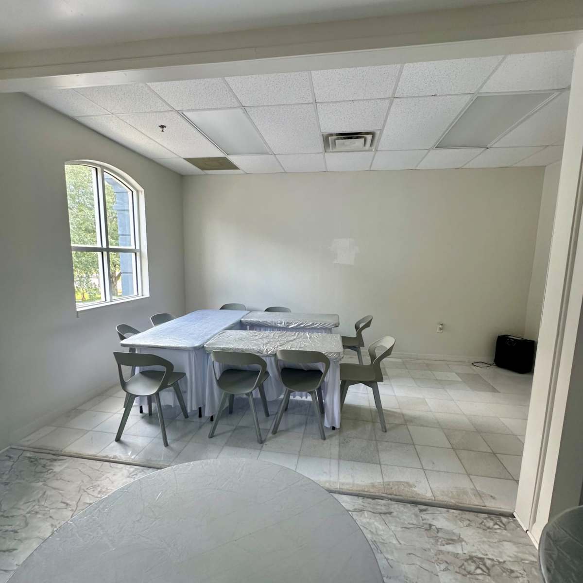 A simple, sparsely furnished room with a rectangular table covered by a white tablecloth and surrounded by eight chairs.