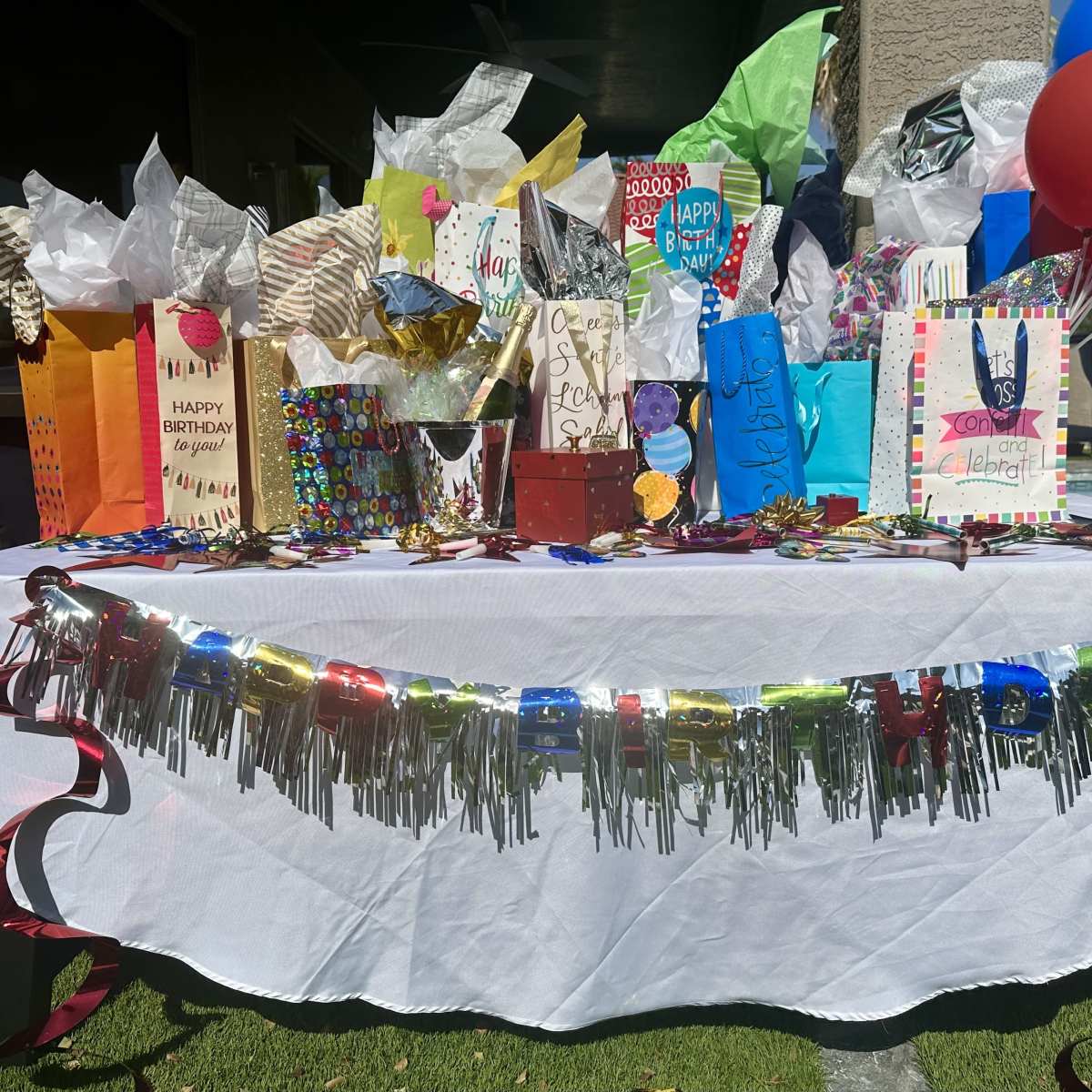 A decorated table is set up outdoors, covered with a white tablecloth and adorned with colorful gift bags, wrapped presents, party hats, and red balloons.